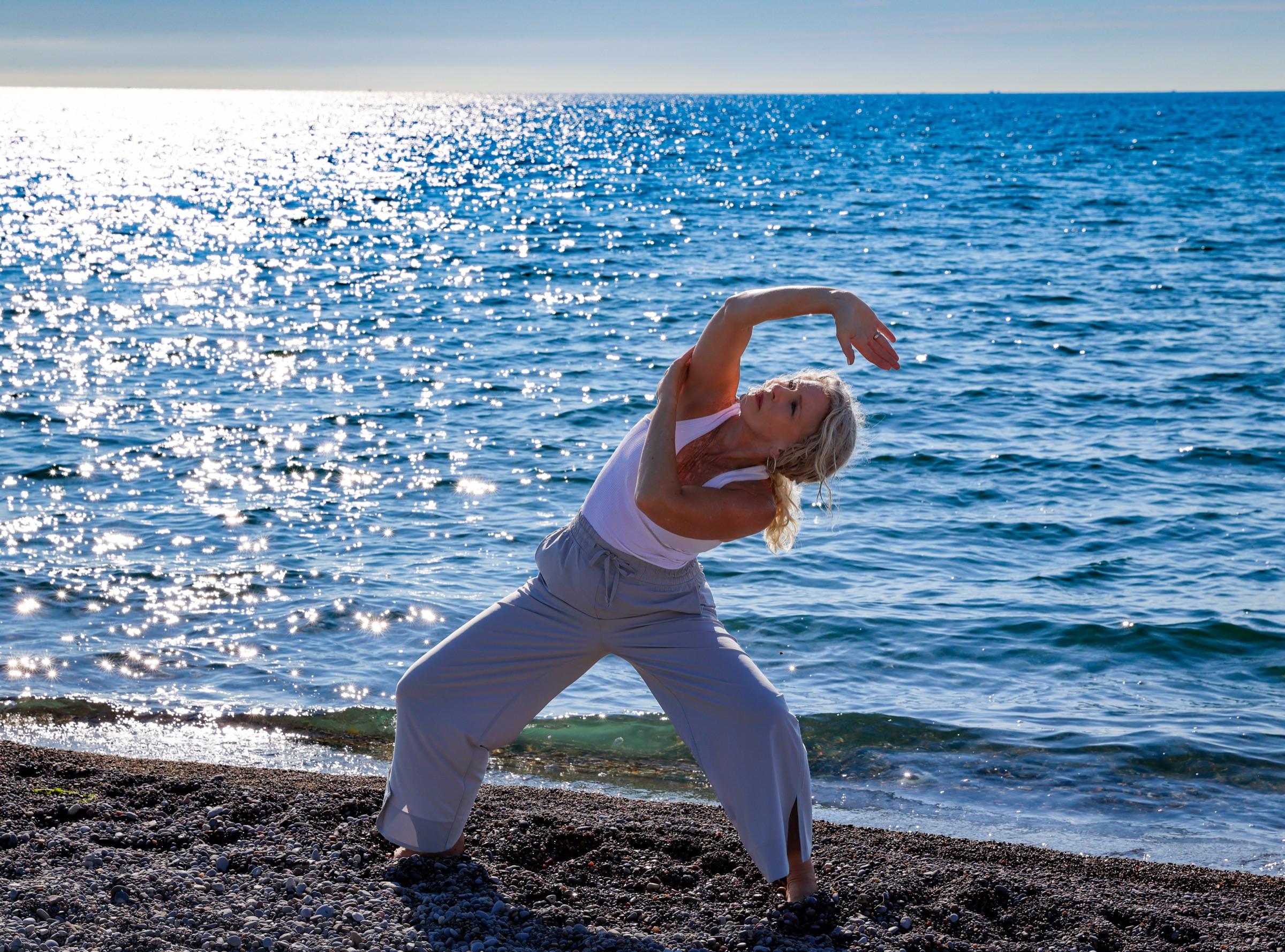 Stephanie Colwell practising Qigong on a lakeshore, arms sweeping into a deep side bend against sparkling blue water